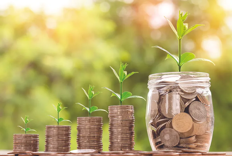 Four stacks of coins and a jar full of coins. The stacks grow in size from left to right. On top of each stack and on top of the jar are plant sprouts increasing in size from left to right.