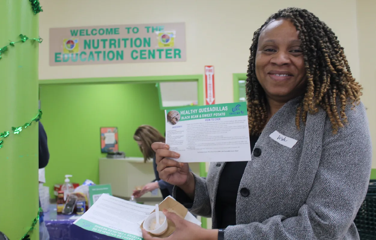 A smiling person stands holding up a flyer. A sign on the wall in the background reads Welcome to the Nutrition Education Center.
