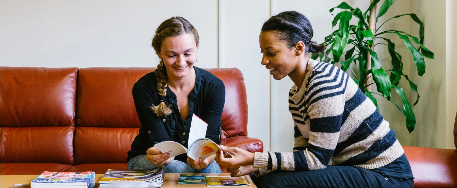 Two women are sitting around a coffee table, looking at a magazine together.