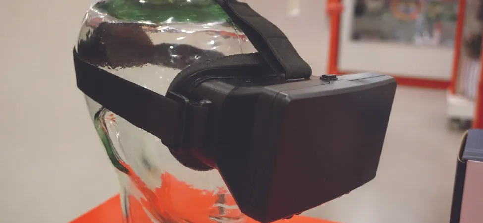 A black virtual reality headset is displayed on a clear glass head model, showcasing its design and strap details in an indoor setting with a blurred background.