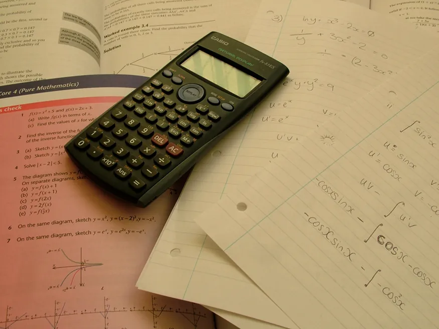 A calculator is placed on open math books and few papers. Problems on trigonometry are solved on one of the papers.