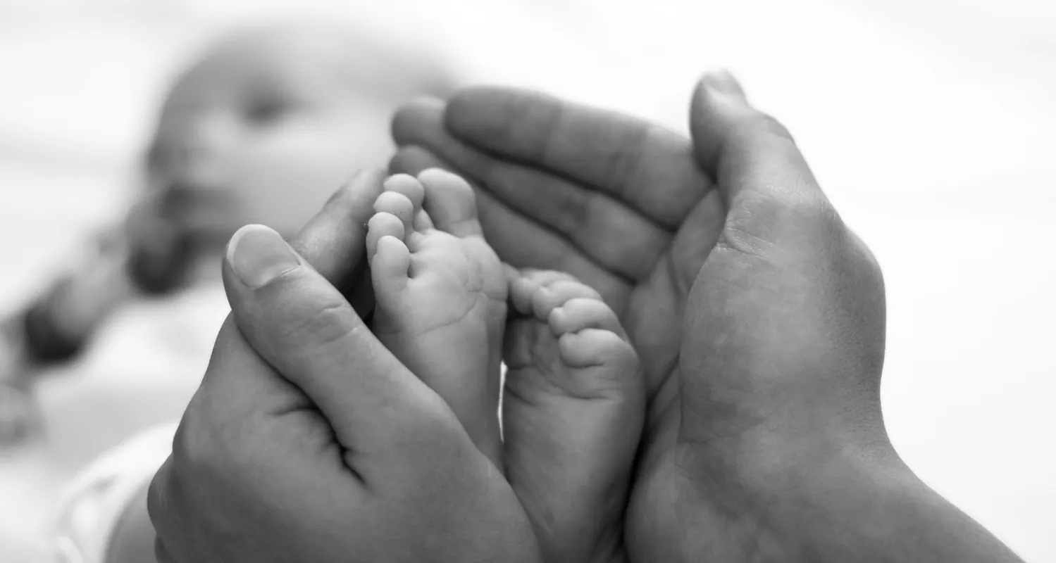 Black and white photo of two adult hands holding the feet of a small baby that is blurry in the background.