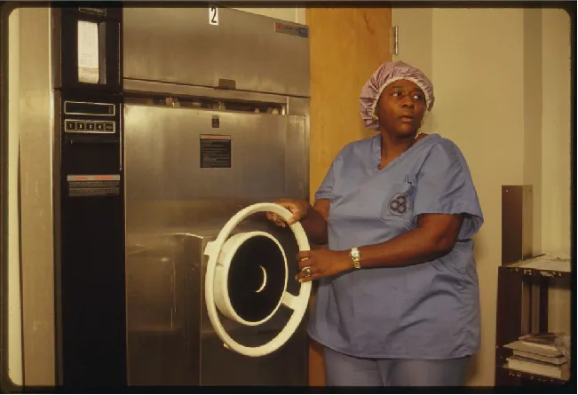 a) A drawing of an autoclave. There is a large metal cylinder with a pressure gauge. An operative valve allows steam from the jacket to the chamber; there is also a safety valve. The main chamber connects to an exhaust valve, a waste line, an ejector valve, a steam supply line and a pressure regulator. B) a photo of an autoclave; a large metal box as tall as the operator standing in front of it.