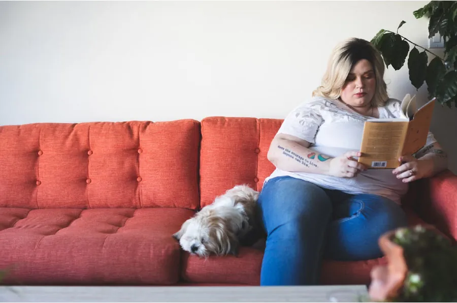 Photo of individual sitting on a couch reading a book with a dog next to them.