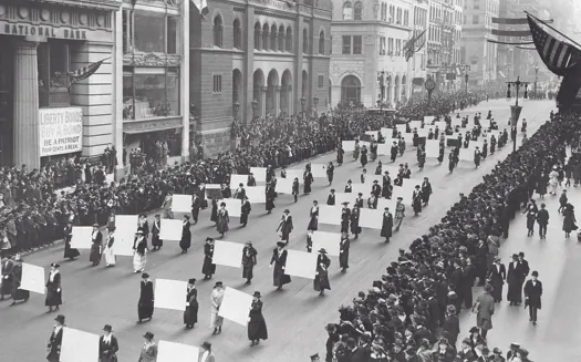 An image of a group of people marching down a street. Several pairs of people are carrying large signs between them. On both sides of the street is a crowd of observers.
