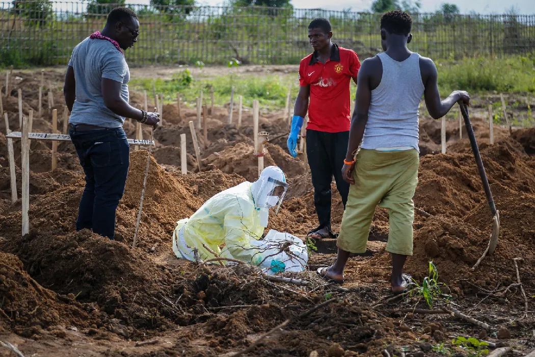 A person wearing full-coverage protective medical clothing kneels in a burial field in which many small wooden crosses stick up from the dirt. Three people in regular clothing stand around this person holding shovels.