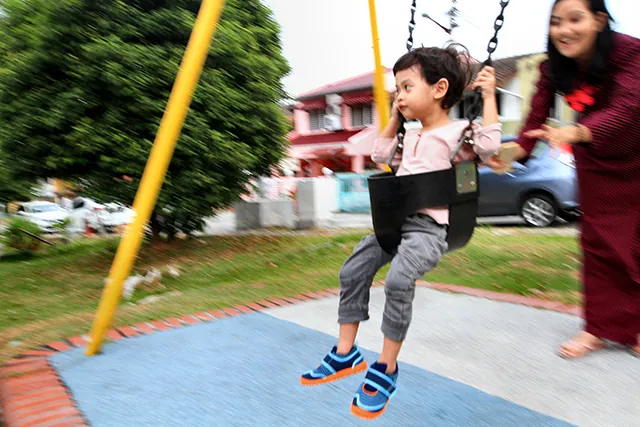 In the figure shown, a small child is seated in a spring swing, tied with a belt at his waist. In the back is his father, who is pushing the swing in the to and fro motion.
