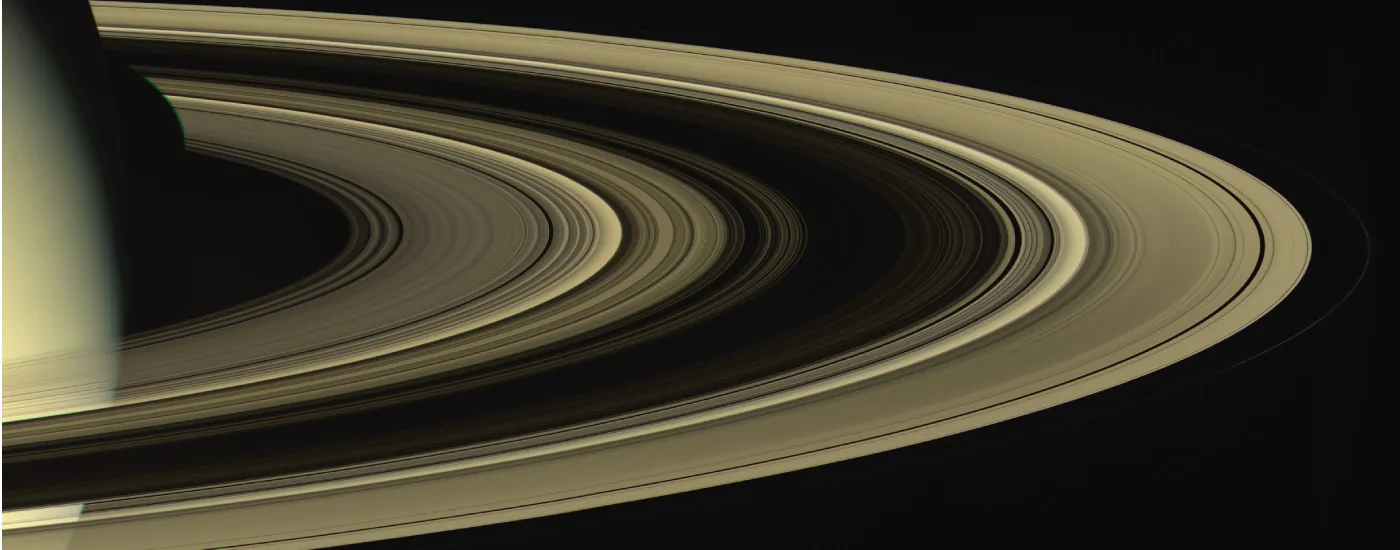 The rings of Saturn are shown from a viewpoint slightly above them, revealing two very wide rings and dozens of smaller ones, with gaps between them. A small portion of Saturn is visible.