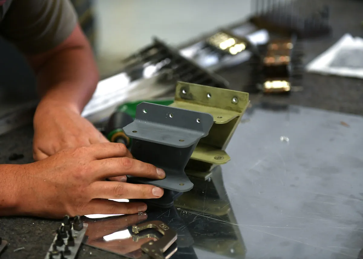 A person holds a metal bracket on a table next to one that appears older but is the same size and shape. The brackets are about 8 to 10 centimeters high and have surfaces at right angles, with premade holes for attachment.