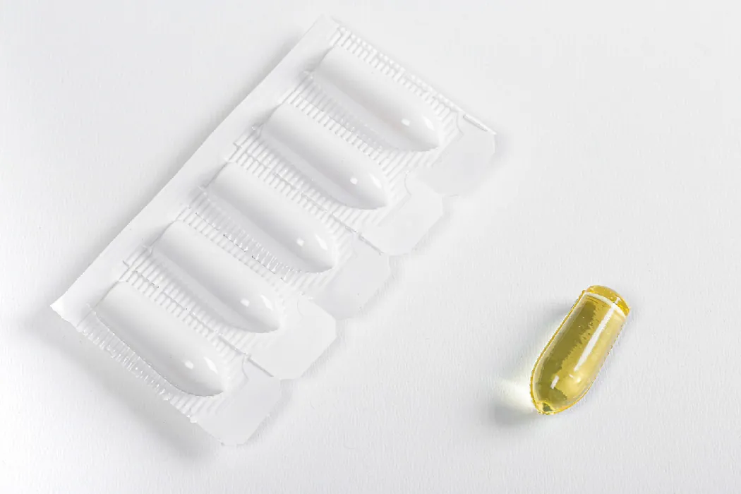 A photograph shows a packet of 5 suppositories still encased in their plastic wrapping. A sixth suppository is unwrapped. The suppositories are narrow, small cylinders with rounded tips.