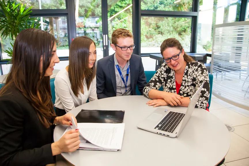 A photograph shows an informal meeting, where 4 people are sitting around a table looking at a laptop.