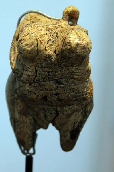 A picture of an old artifact is shown on a blue background. It is rectangular and colored black and yellow. A small loop is shown at the top right. It has two bumps on the front toward the top and lines running horizontally across the middle with two short legs coming out of the bottom. It is shown displayed on a rod anchored to the left projection at the bottom.