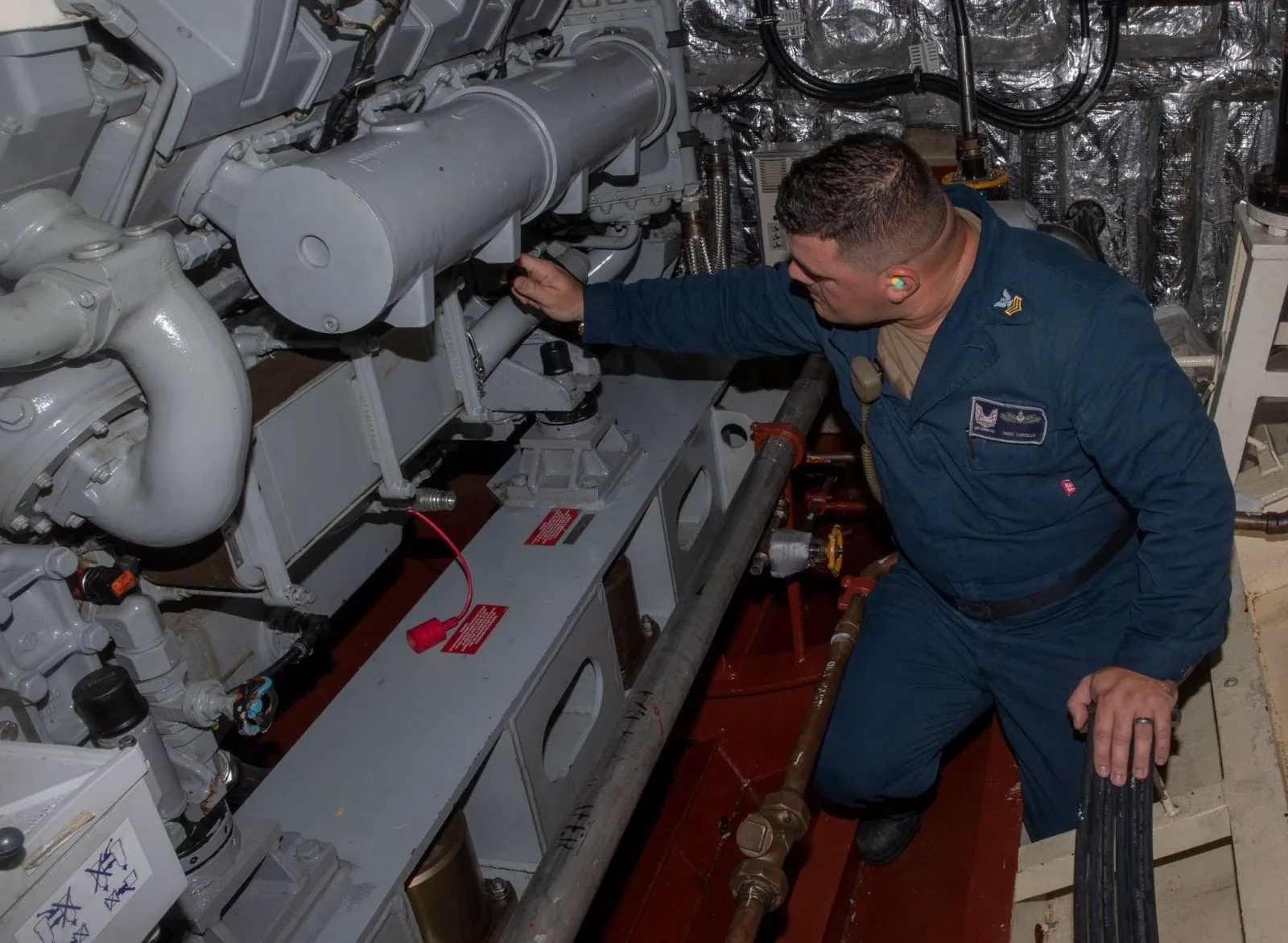 A person crouches in an engine room with an array of large pipes, junctions, gaskets, and cables. The person reaches toward a component on one of the large conduits. 