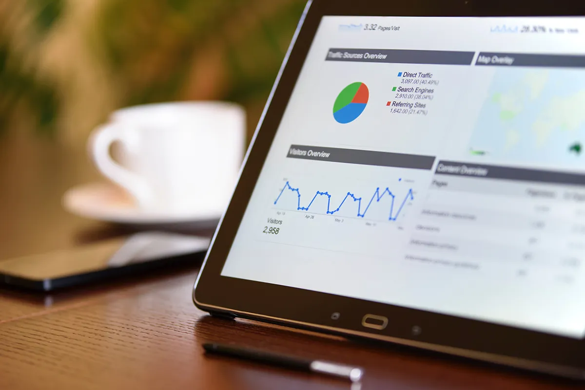 A tablet propped up on a desk with a data dashboard in view. The dashboard has a pie chart, a graph, a map, and additional rows of data. A coffee cup and phone are out of focus in the background.