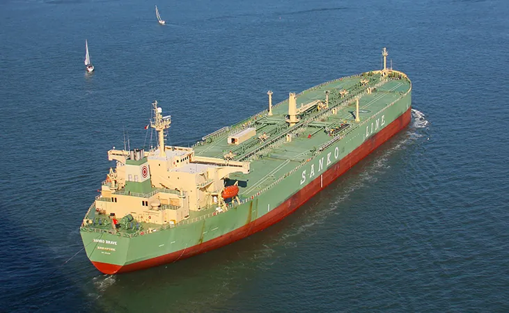 Se muestra una foto de un supertanquero en el agua. Hay dos barcos mucho más pequeños con velas en la distancia.