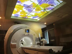 A modern MRI machine in a medical room, designed with a comforting, illuminated ceiling panel featuring vibrant yellow and purple flowers to soothe patients during scans.