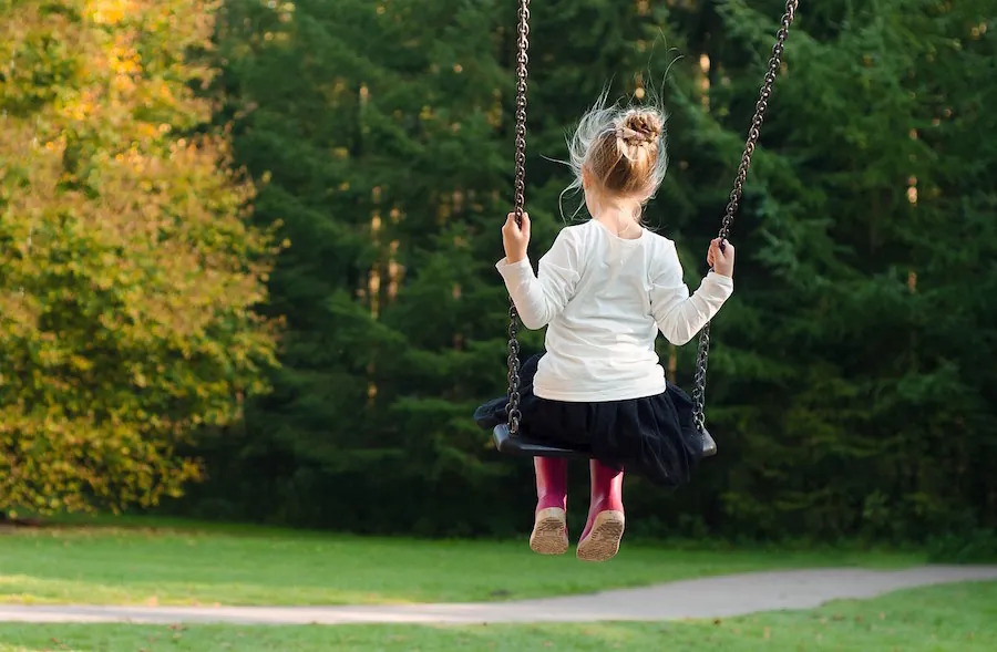 A young girl with her hair tied up in a bun swings high on a playground swing, facing away from the camera towards the lush green trees and a winding path.