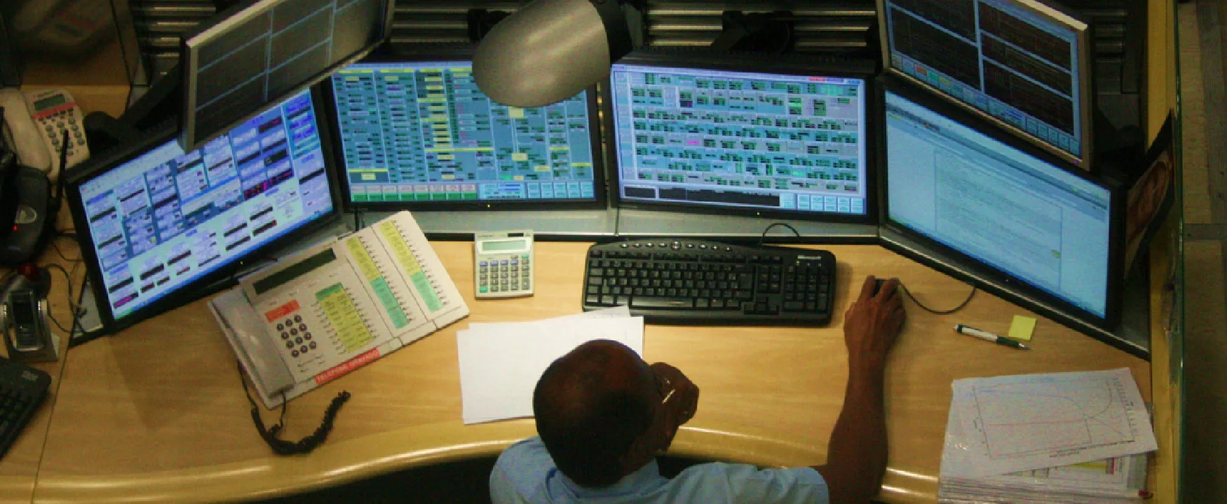 Overhead view of a person intensely working at a multi-monitor workstation, surrounded by screens displaying complex data and charts, a phone, calculator, and keyboard.