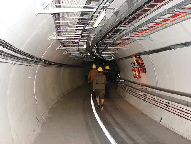 Two photos are shown and labeled “a” and “b.” Photo a shows an aerial view of the Large Hadron Collider. Photo b shows a tunnel of concrete with rails on the ground and tubes and wires running along the wall. Two people walk along the tunnel.