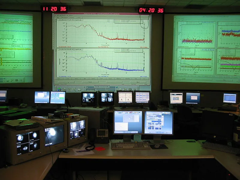 This figure shows a windowless room full of desks and computer screens and with three large screens on the wall upon which are projected a lot of technical graphs.