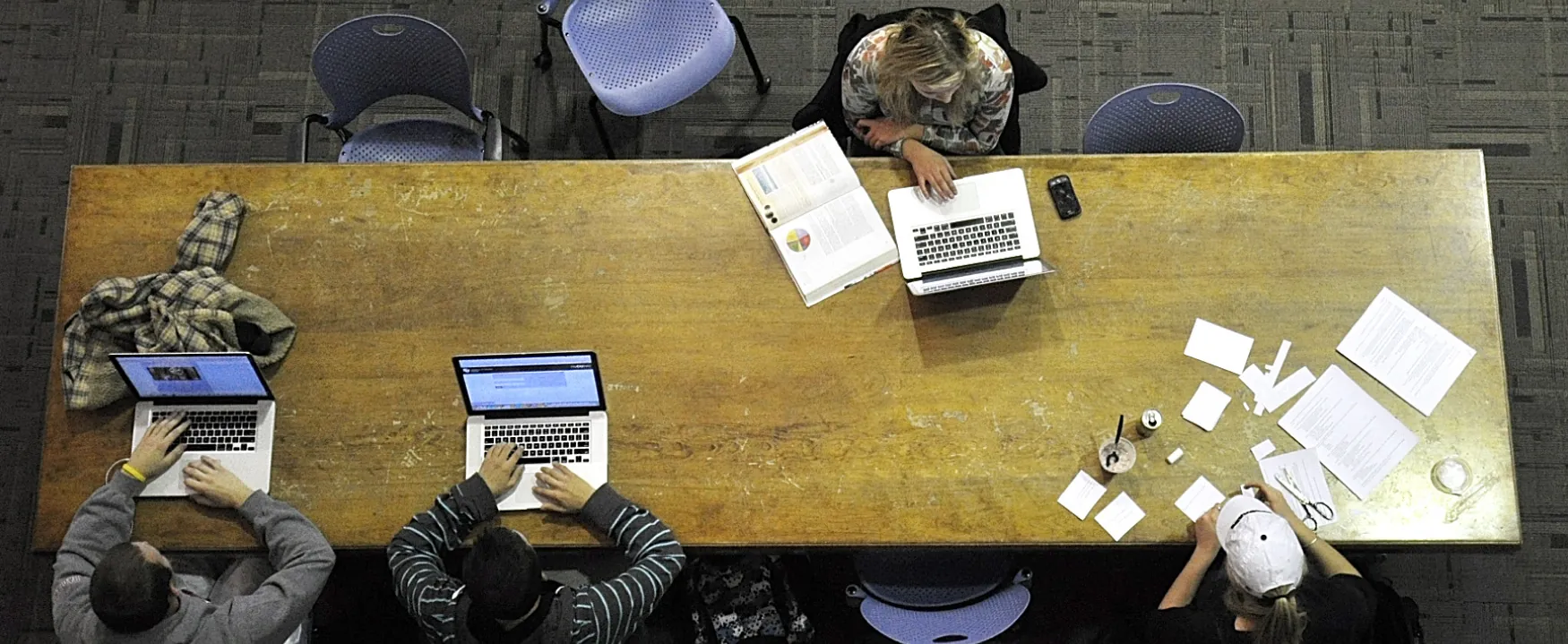 Four people sit at a long table. Two have computers, while another has a computer and what appears to be a textbook. The fourth person has a number of papers and is cutting them into smaller pieces.