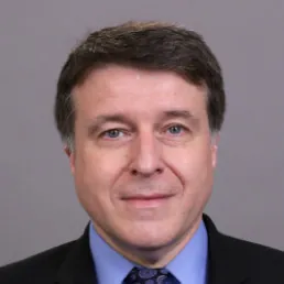 Headshot of a smiling man in professional attire, featuring a dark suit and blue shirt with a patterned tie, on a gray backdrop.